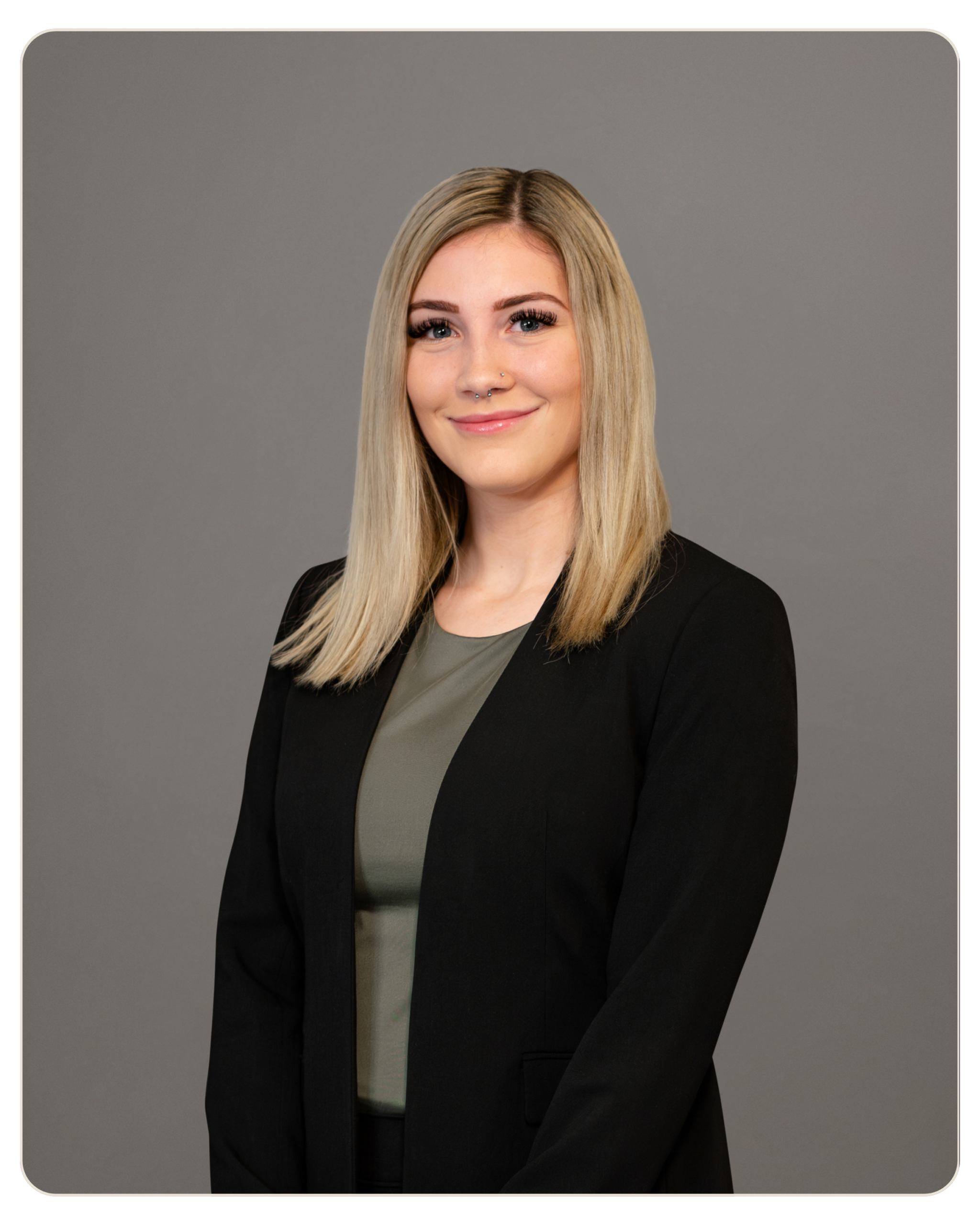Nicole Laska Smiling young woman with blonde hair in a black blazer, posing for a corporate headshot against a gray background.