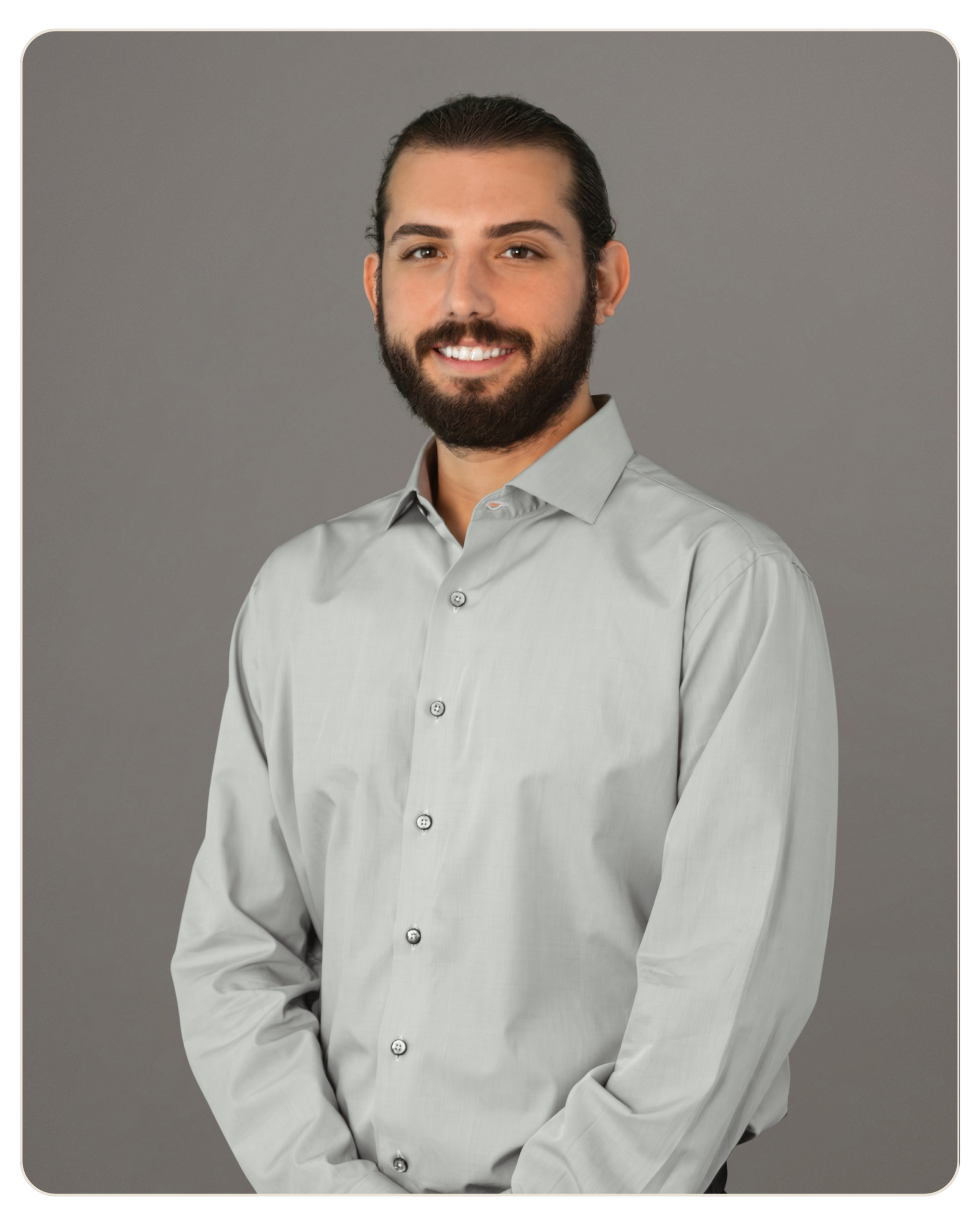 Flyland - Trost - Robert Mazo Portrait of a man with dark hair and a full beard, smiling slightly, wearing a light gray button-down shirt against a neutral gray backdrop.